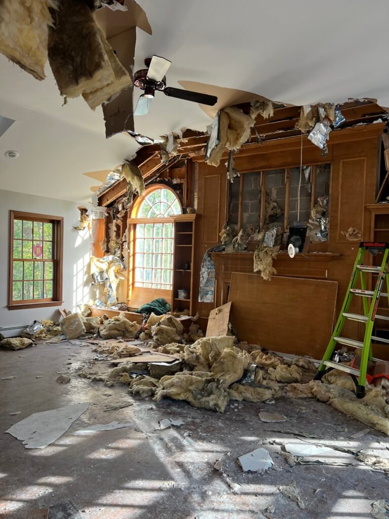 Room with significant ceiling damage, debris, insulation, and wood scattered on the floor after a storm; ladder near wooden shelves and large arched window—ideal for Storm Damage Restoration in Garnet Valley PA.