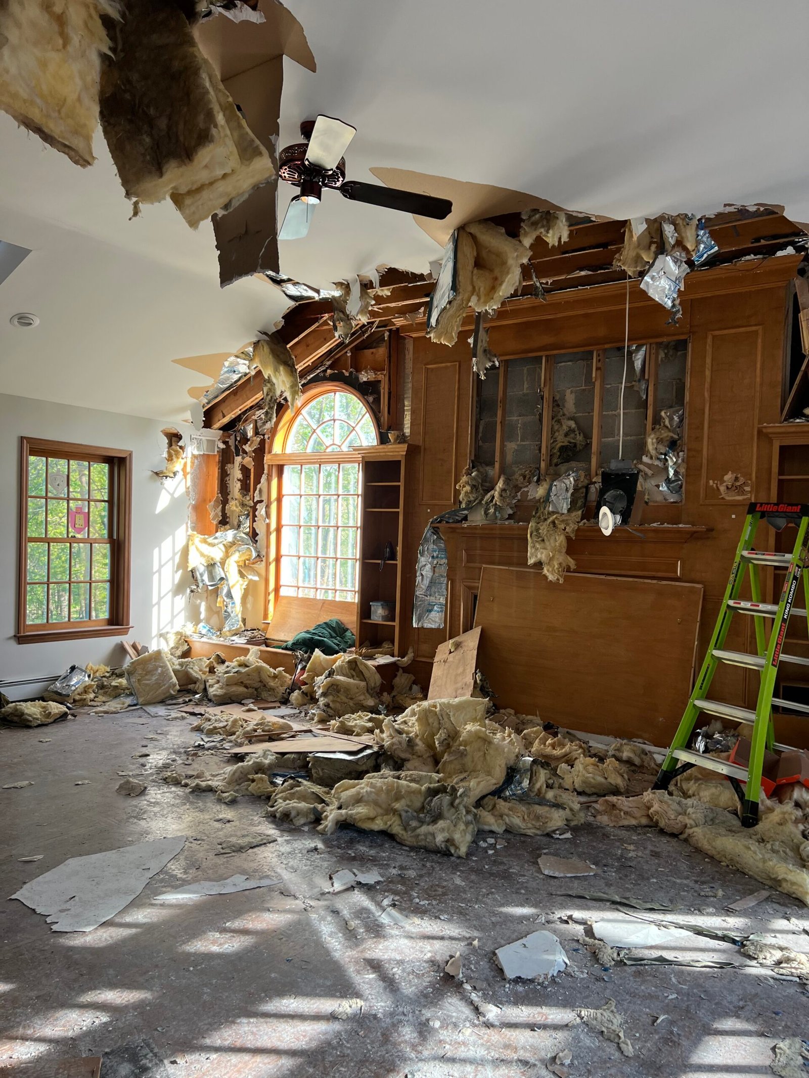 Room with significant ceiling damage, debris, insulation, and wood scattered on the floor after a storm; ladder near wooden shelves and large arched window—ideal for Storm Damage Restoration in Garnet Valley PA.