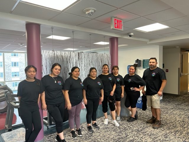 Seven women and one man stand in a row indoors at a Garnet Valley gym, all wearing black t-shirts and casual pants, posing for a group photo near gym equipment.