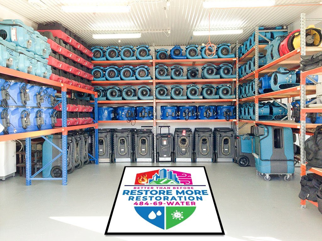 Industrial restoration equipment for water damage restoration neatly organized on shelves in a warehouse, with a large Restore More Restoration logo and contact number displayed on the floor.