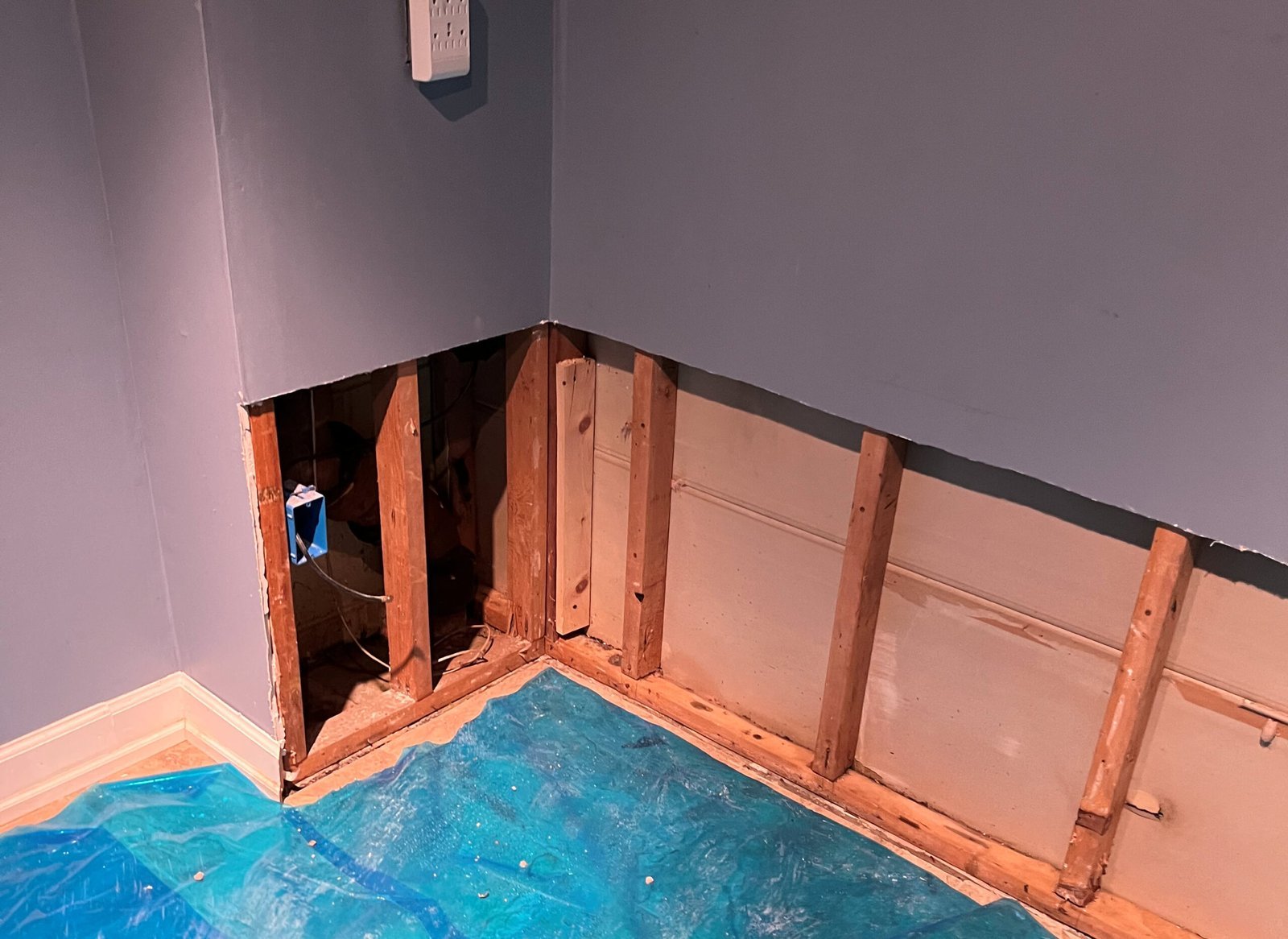 A section of drywall is removed from a corner wall, exposing wooden studs and wiring; a blue tarp covers part of the floor during water damage restoration.